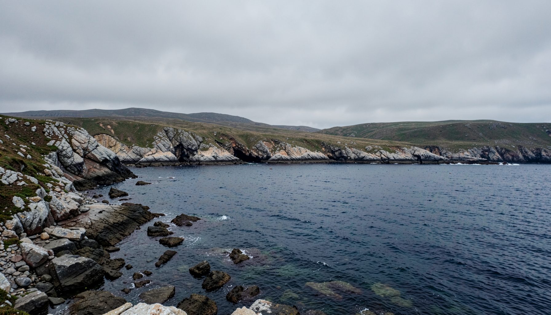 Atmospheric wide landscape of a rocky northern coastline under a grey overcast sky, dark blue-grey water meeting pale stone, moody and solitary