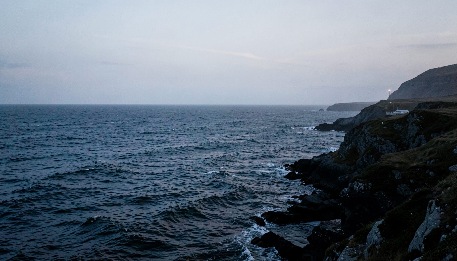 A windswept northern coastline at dusk, deep grey-blue sea meeting a pale sky, evoking the atmospheric setting of Nordic crime fiction